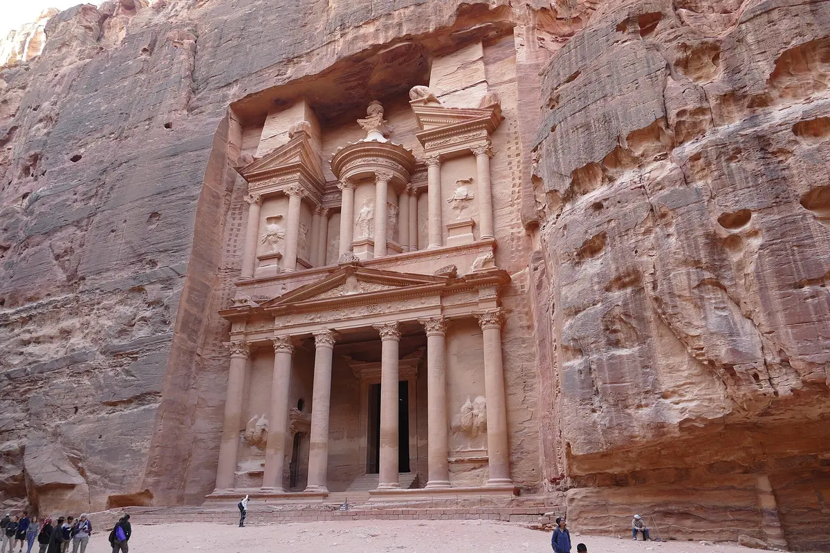 The full carved facade of the Treasury at Petra, with its columns and upper tholos cut into a rose-coloured cliff, and a few visitors on the sand in front